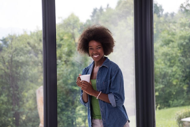 Hermosa joven afroamericana tomando café y mirando a través de una ventana en su casa de lujo