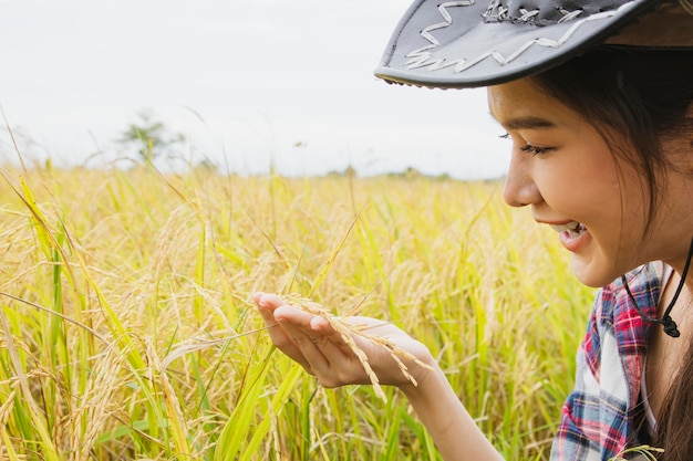 Hermosa joven agricultora asiática mira la planta de arroz dorado en su mano con una sonrisa y orgullo.