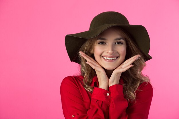 Hermosa joven alegre con una camisa roja y un sombrero en una pared rosa