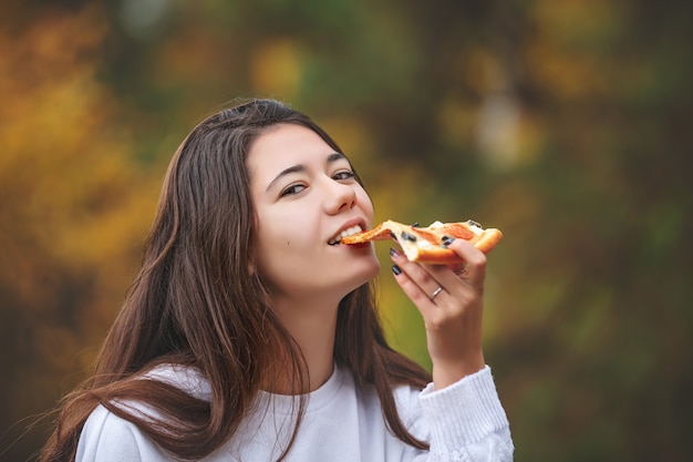 Hermosa joven alegre comiendo pizza mientras disfruta del sabor en la naturaleza