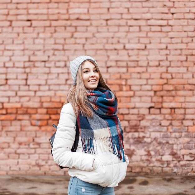 Hermosa joven alegre con una dulce sonrisa en una chaqueta blanca y una bufanda colorida con una bolsa negra cerca de la pared de ladrillo