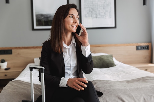 hermosa joven alegre mujer de negocios feliz en ropa formal en el interior en casa con maleta hablando por teléfono móvil.