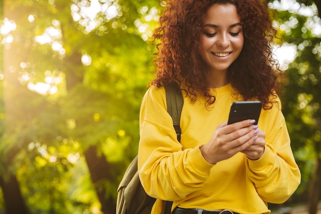 Hermosa joven alegre pasar buen rato en el parque, enviando mensajes de texto en el teléfono móvil