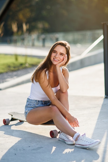 Hermosa joven alegre se sienta en patineta en el espacio urbano al amanecer.