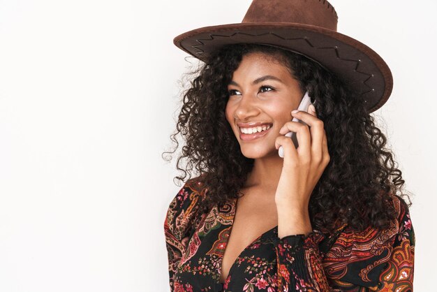 Hermosa joven alegre con sombrero de vaquero que se encuentran aisladas sobre una pared blanca, hablando por teléfono móvil