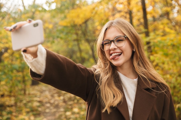 Hermosa joven alegre vistiendo abrigo caminando en el parque de otoño, tomando un selfie