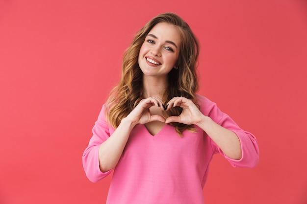 Hermosa joven alegre vistiendo ropa casual que se encuentran aisladas sobre la pared rosa, mostrando el gesto del corazón