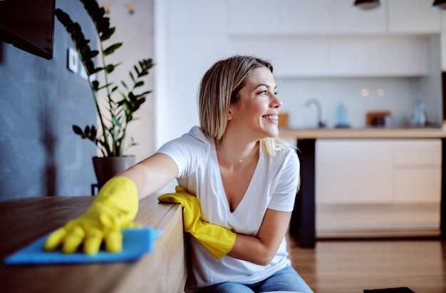 Hermosa joven ama de casa rubia caucásica con guantes de goma en las manos sentado en el suelo en la sala de estar y desempolvando estante. Salón interior.