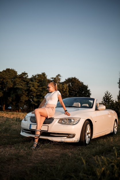 Foto una hermosa joven de apariencia europea en el rostro de las gafas de sol, se encuentra junto a su convertible blanco. mujer de negocios acertada y su coche blanco.