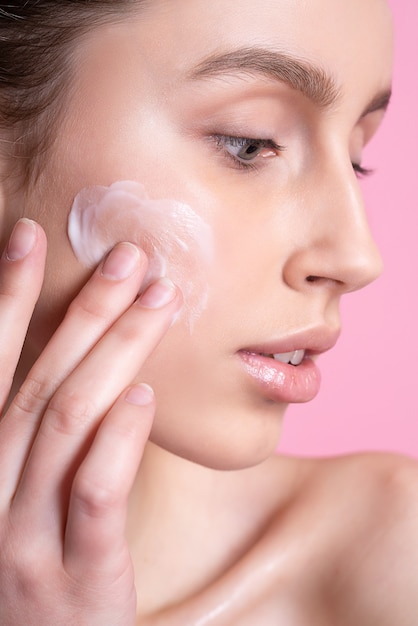 Hermosa joven aplicando crema en el rostro en rosa