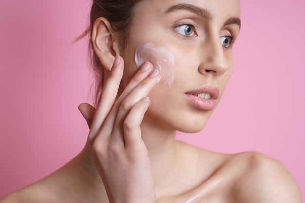 Hermosa joven aplicando crema en el rostro en rosa