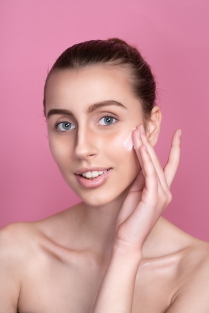 Hermosa joven aplicando crema en el rostro en rosa