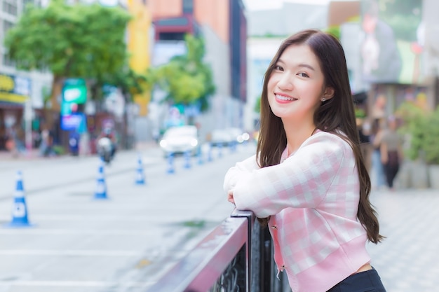Hermosa joven asiática con un abrigo de rayas rosa y blanco está de pie al aire libre urbano con la ciudad y la carretera como fondo.