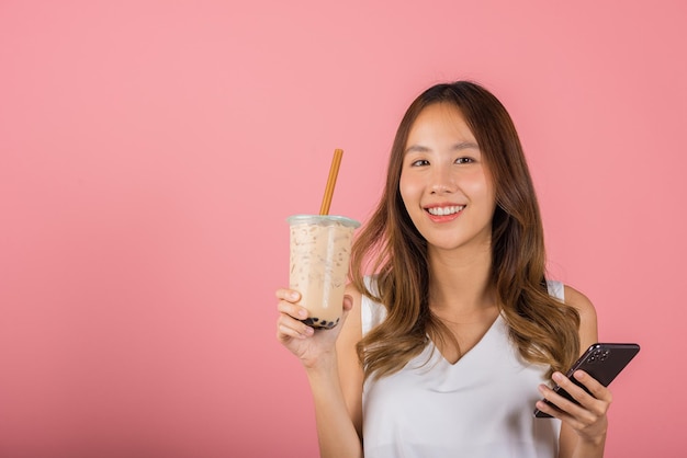 Hermosa joven asiática bebiendo té de leche con burbujas de perla de tapioca con sabor a azúcar moreno, retrato sonriente femenino sosteniendo teléfono inteligente, foto de estudio aislada en fondo rosa, concepto de bebida de leche