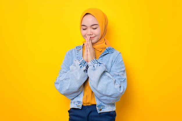 Hermosa joven asiática con chaqueta de jeans mantiene las palmas juntas en un gesto de oración aislado en un fondo amarillo