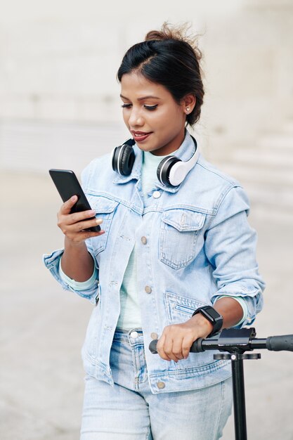 Hermosa joven asiática en chaqueta de mezclilla de pie al aire libre, apoyado en el manillar del scooter eléctrico y leyendo un mensaje de texto