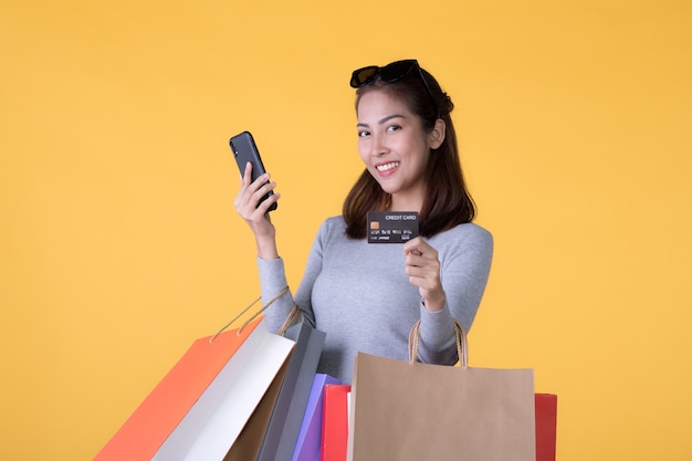 Hermosa joven asiática con coloridas bolsas de la compra con teléfono inteligente y tarjeta de crédito aislado en la pared amarilla