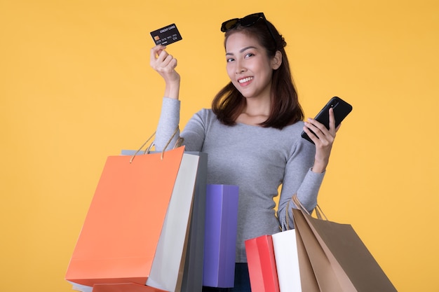 Hermosa joven asiática con coloridas bolsas de la compra con teléfono inteligente y tarjeta de crédito aislado en la pared amarilla