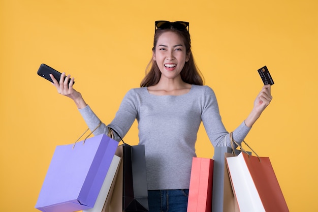 Hermosa joven asiática con coloridas bolsas de la compra con teléfono inteligente y tarjeta de crédito aislado en la pared amarilla