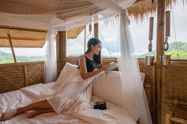 Hermosa joven asiática descansando y leyendo un libro en la cama en la choza de paja entre la montaña de vacaciones