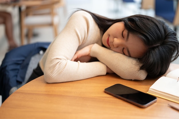 Una hermosa joven asiática se está quedando dormida en una mesa en una biblioteca o cafetería