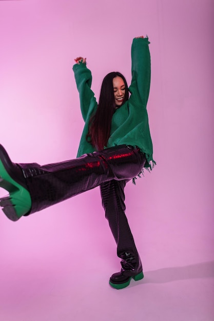 Hermosa joven asiática feliz con ropa de moda con un suéter verde rasgado, pantalones de cuero y botas, se divierte y se divierte con un fondo rosa