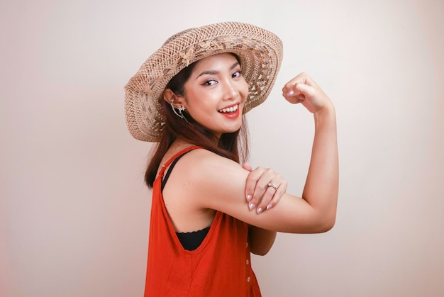 Hermosa joven asiática fuerte con vestido naranja y sombrero de paja mostrando bíceps y sonriente concepto fuerte de niña indonesia