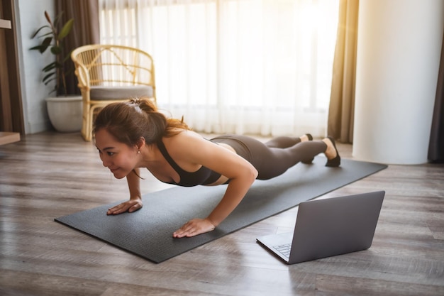 Una hermosa joven asiática haciendo flexiones en la colchoneta de entrenamiento mientras ve tutoriales de entrenamiento en línea en la computadora portátil en casa