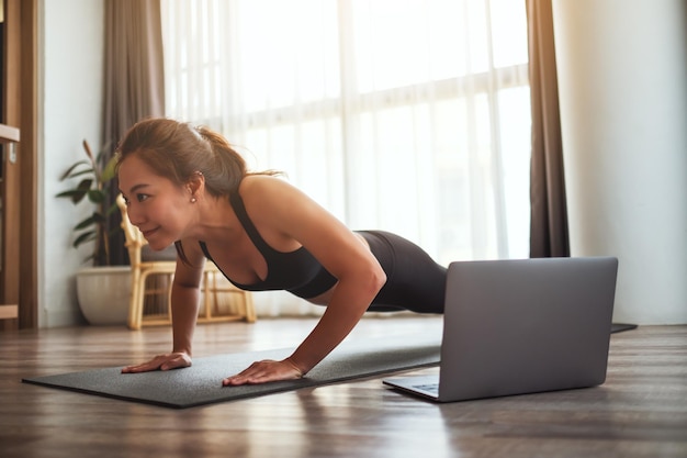 Una hermosa joven asiática haciendo flexiones en la colchoneta de entrenamiento mientras ve tutoriales de entrenamiento en línea en la computadora portátil en casa