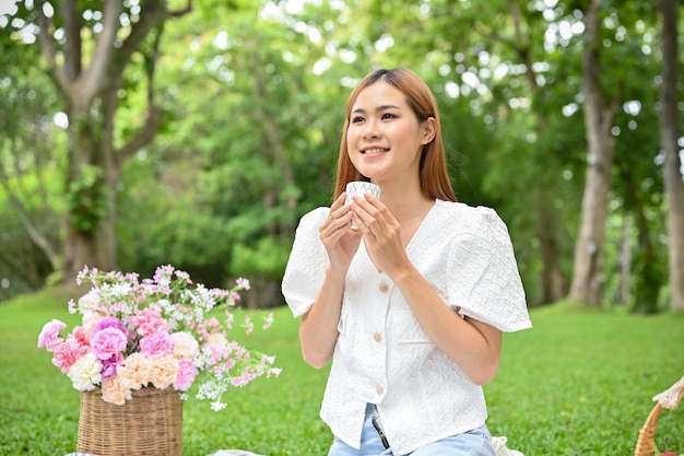 Hermosa joven asiática haciendo un picnic en el parque verde bebiendo un té caliente