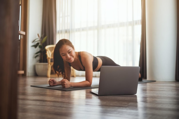 Una hermosa joven asiática haciendo plancha en la estera de entrenamiento mientras ve tutoriales de entrenamiento en línea en la computadora portátil en casa