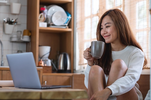 Una hermosa joven asiática independiente tomando café y usando una computadora portátil para trabajar en línea en casa
