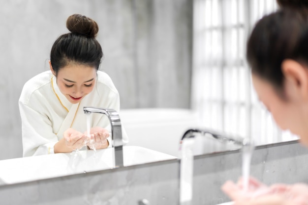 Hermosa joven asiática lavando la cara limpia con agua y sonriendo frente al espejo en el baño. Belleza y spa. Perfecta piel fresca