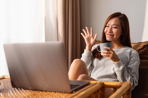 Una hermosa joven asiática que usa una computadora portátil para una videollamada mientras se sienta y toma café en casa