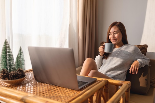 Una hermosa joven asiática que usa y trabaja en una computadora portátil mientras toma café en casa