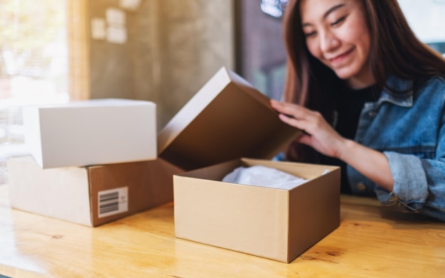 Una hermosa joven asiática recibiendo y abriendo una caja de paquetería postal en casa para la entrega y el concepto de compras en línea