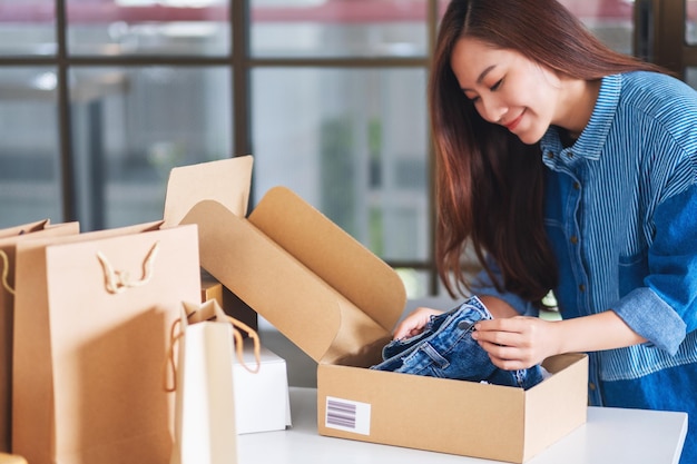 Una hermosa joven asiática recibiendo y abriendo una caja de paquetes postales de ropa en casa para la entrega y el concepto de compras en línea