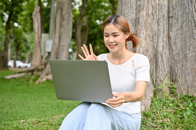 Hermosa joven asiática se relaja mientras se sienta debajo del árbol usando su computadora portátil para hacer una videollamada