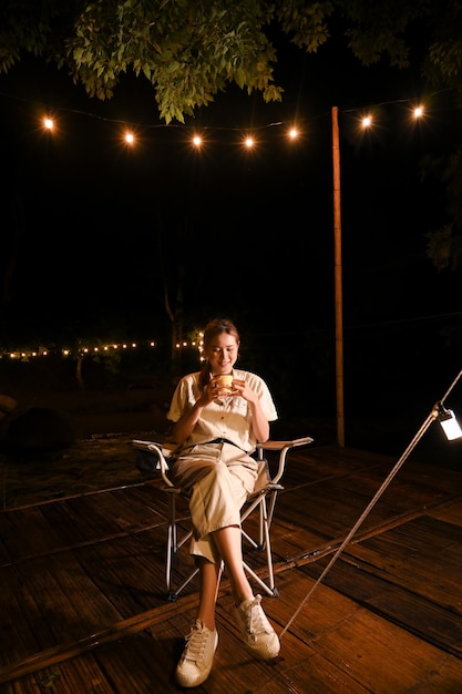 Hermosa joven asiática relajándose bebiendo un chocolate caliente en el hermoso camping por la noche