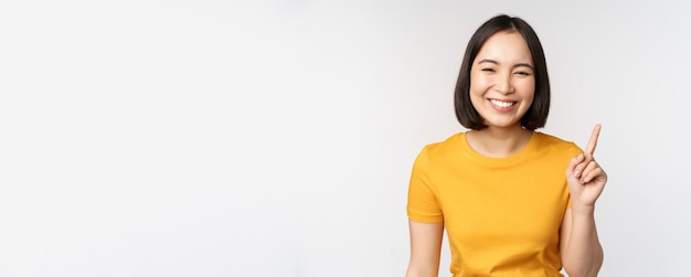 Hermosa joven asiática señalando con el dedo sonriendo y mirando divertida a la cámara que muestra un anuncio publicitario en el fondo blanco superior
