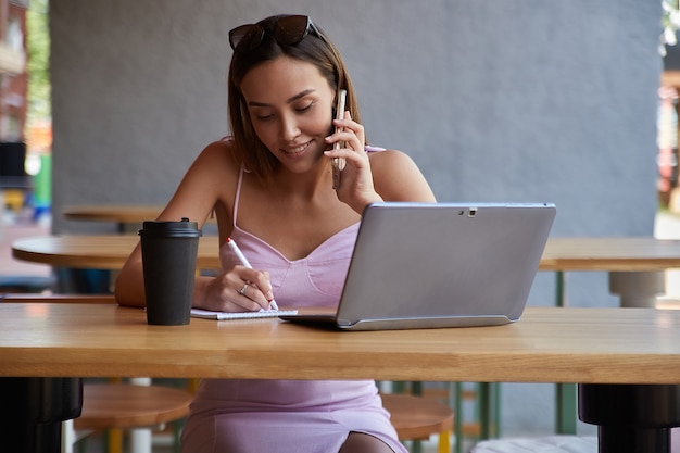 Hermosa joven asiática sentada con el portátil en el café llamando por teléfono inteligente