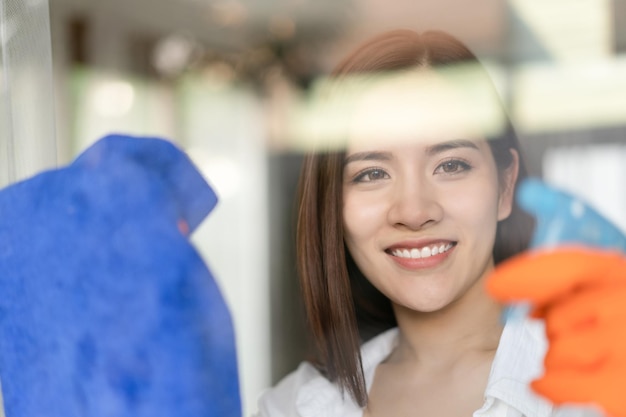 Hermosa joven asiática sonriente limpia la ventana con un trapo Concepto de limpieza y limpieza