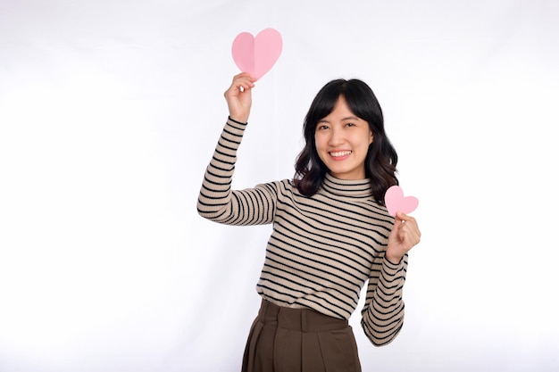 Hermosa joven asiática sosteniendo un corazón de papel mientras está de pie contra el fondo blanco Hermosa joven asiática con corazón de papel