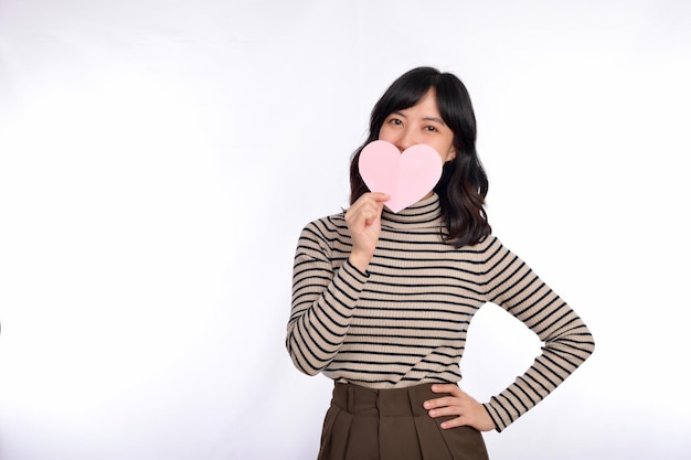 Hermosa joven asiática sosteniendo un corazón de papel mientras está de pie contra el fondo blanco Hermosa joven asiática con corazón de papel