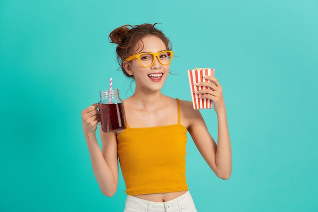 Hermosa joven asiática sosteniendo palomitas de maíz y bebiendo refrescos sobre azul.