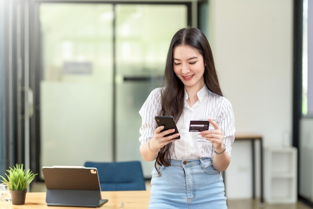 Hermosa joven asiática sosteniendo un teléfono inteligente y usando tarjeta de crédito para comprar en línea.