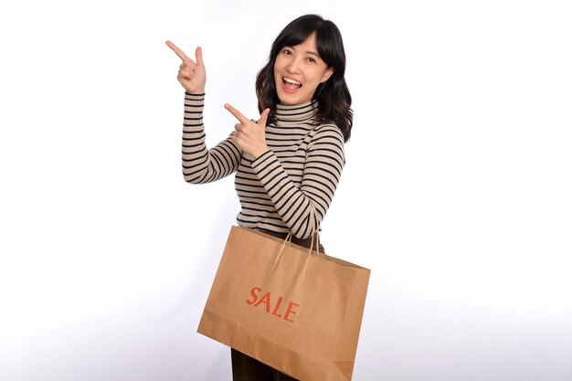 Hermosa joven asiática en tela de suéter apuntando hacia arriba para copiar espacio y mirando a la cámara con cara de sonrisa y bolsa de papel de compras de venta aislada en fondo blanco