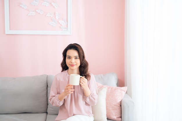 Hermosa joven asiática tomando café y relajándose en el sofá.