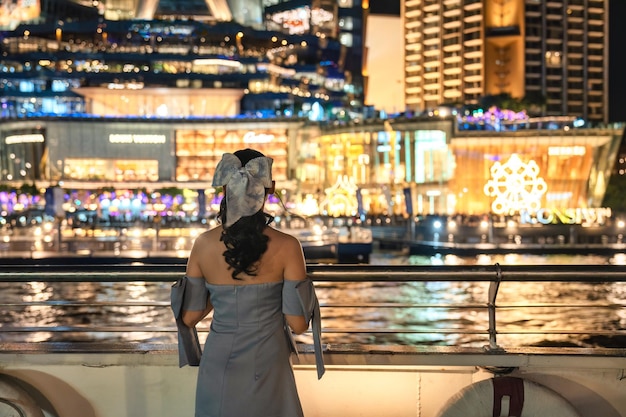 Hermosa joven asiática vestida mirando los grandes almacenes iluminados que brillan intensamente en un evento festivo en la orilla del río en la noche Crucero con cena navegando y haciendo turismo en el río Chao Phraya