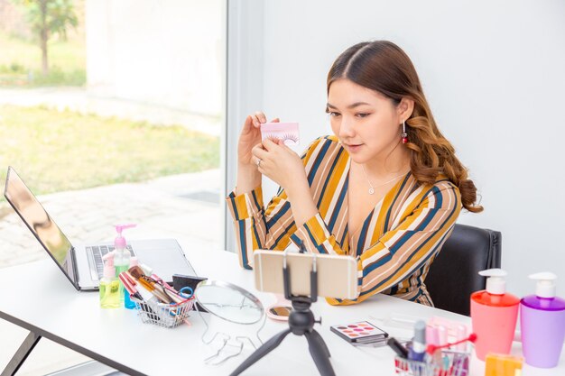 Hermosa joven asiática, vlogger, mostrando extensión de pestañas mientras revisa productos de belleza en un video blog